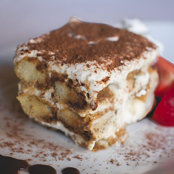 A close-up of a slice of tiramisu with cocoa powder, whipped cream, and strawberries.