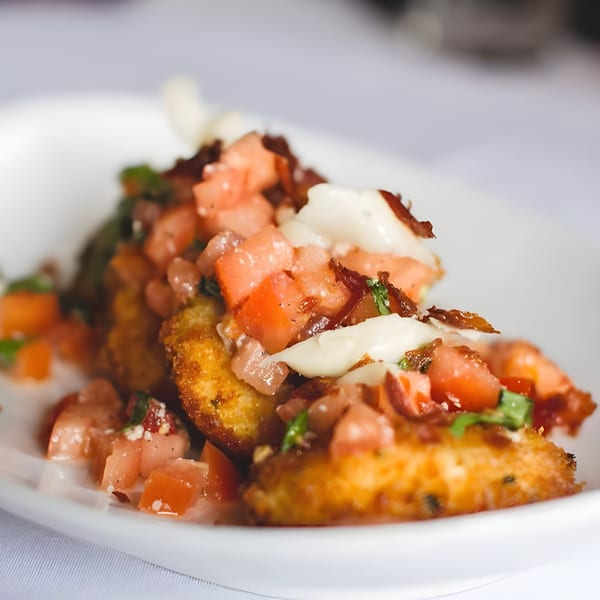 Close-up of golden-fried Italian appetizers topped with diced tomatoes, bacon, and creamy white sauce.